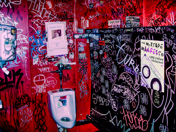 Red grafitti bathroom with stickers on the urinal. A poster of the book The Mixtape Manifesto: A Pop Culture Confessional